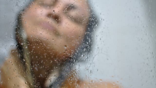 Tummahiuksinen nainen suihkussa näkyvillä ainoastaan kasvot ja olkapää sekä suihkulasin vesipisarat
